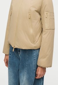 Veste matelassée beige à fermeture éclair, avec poches zippées à l'avant et sur la manche gauche, portée avec un jean denim bleu.