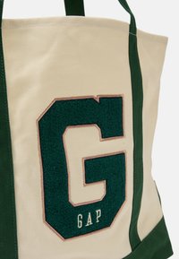 Platnena torba s zelenim detaljima i teksturiranim zelenim logotipom "G". Krem pozadina s "GAP" u bijeloj boji ispod. Dvostruki ručici za nošenje.