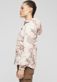 Lätt, pastellfärgad kamouflagejacka med en figurnära huva, elastiska muddar och en dragkedja framtill. Materialet är slätt med en matt yta.