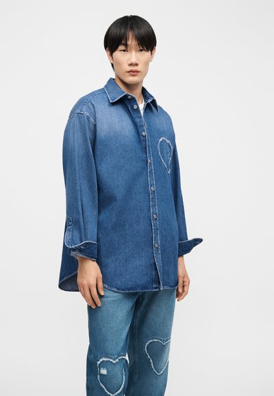 Jeune personne portant une chemise en denim bleu assortie et un jean avec des patchs en forme de cœur sur la poitrine et les genoux, debout devant un fond blanc.