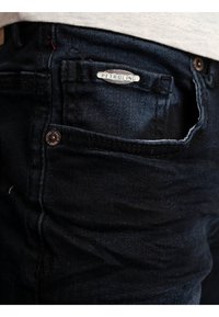 Jeans de mezclilla oscura con un logo de PETROLIND en el bolsillo delantero, acentos de botón de cobre y una tela texturizada con un ligero brillo.