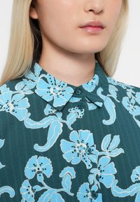 Nærbilde av person som har på seg en teal skjorte med lys blå blomsterdesign og knappet krage, som viser lepper og nedre del av ansiktet.