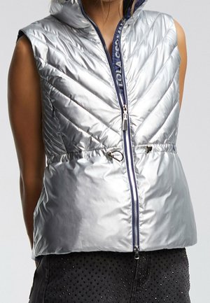 Mouwloos zilveren puffer-vest met capuchon, ritssluiting aan de voorkant en verstelbaar trekkoord in de taille, gedragen over donkere geperforeerde broek.