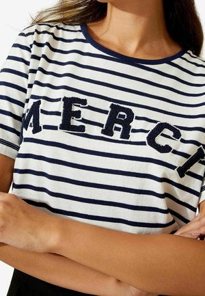 Chemise rayée en blanc et marine avec un col rond. Le mot "MERCI" est présenté en tissu texturé marine. Manches courtes, coupe décontractée.