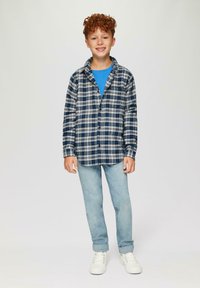Geruite blouse met knopen in marineblauw en wit, gecombineerd met een blauwe T-shirt en lichtblauwe jeans. Heeft krullend haar en witte sneakers.