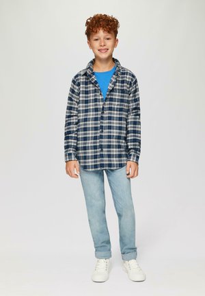Geruite blouse met knopen in marineblauw en wit, gecombineerd met een blauwe T-shirt en lichtblauwe jeans. Heeft krullend haar en witte sneakers.