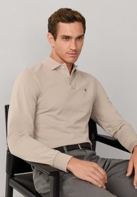 Polo beige à manches longues avec col, arborant un logo discret sur la poitrine, porté avec un pantalon gris, assis sur une chaise noire.