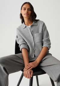 Femme assise sur une chaise, portant un cardigan gris à côtes avec une grande poche et un pantalon large assorti, les mains reposant sur ses genoux.