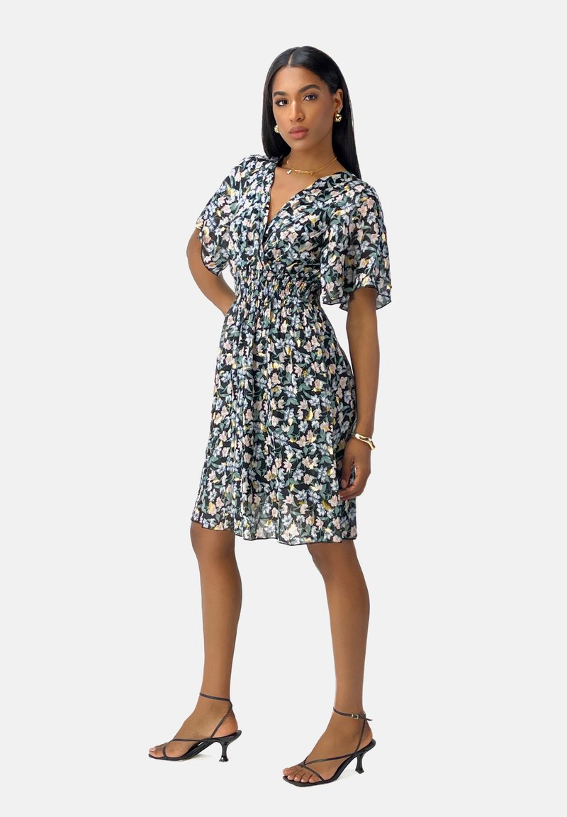 Vestido floral con mangas cortas y ligeras, cintura ceñida y dobladillo a la altura de la rodilla en una tela oscura con patrones en blanco, rosa y verde. Sandalias negras de tiras.