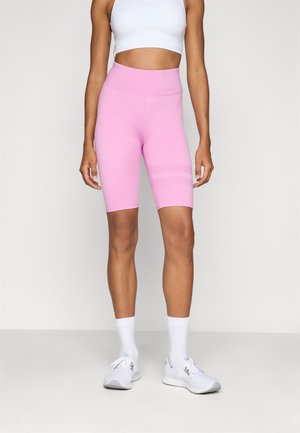 Mujer con pantalones cortos de ciclista rosa de cintura alta, top deportivo blanco corto, calcetines blancos y zapatillas blancas, de pie frente a un fondo liso.