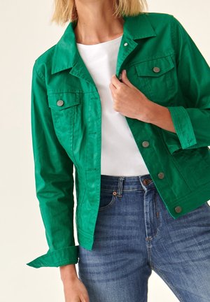 Person trägt eine grüne, geknöpfte Jacke über einem weißen Hemd, kombiniert mit blauen Jeans, Hand passt die Jacke nahe der Brust an.