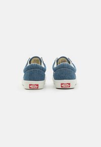 Saboți din denim albastru, fără șireturi, cu tălpi din cauciuc alb și logo roșu Vans pe călcâi. Au o textură netedă și un design cu vârf rotunjit.