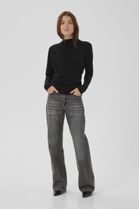 Žena stoji s rukama u džepovima noseći crnu majicu dugih rukava i izblijedjele široke sive traperice na jednostavnoj svijetloj pozadini.