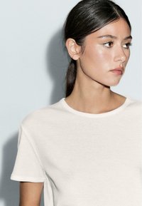 Hvid crewneck t-shirt, korte ærmer, blød bomuldsstruktur, tætsiddende design. Modelens profil synlig mod en lys baggrund.