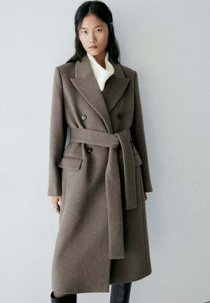 Une femme pose devant un mur gris uni, vêtue d’un long manteau croisé taupe à ceinture nouée par-dessus un pull blanc.