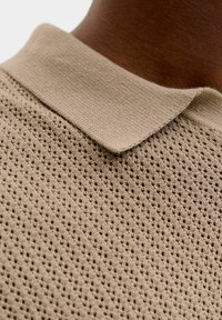 Polo en maille beige avec un motif texturé et perforé porté sur une peau foncée, montrant un gros plan du col et de la partie supérieure de la poitrine.