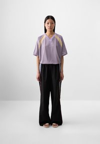 Une jeune femme aux longs cheveux noirs se tient face à l'objectif, portant un t-shirt sportif lavande et un pantalon noir ample, sur un fond uni.