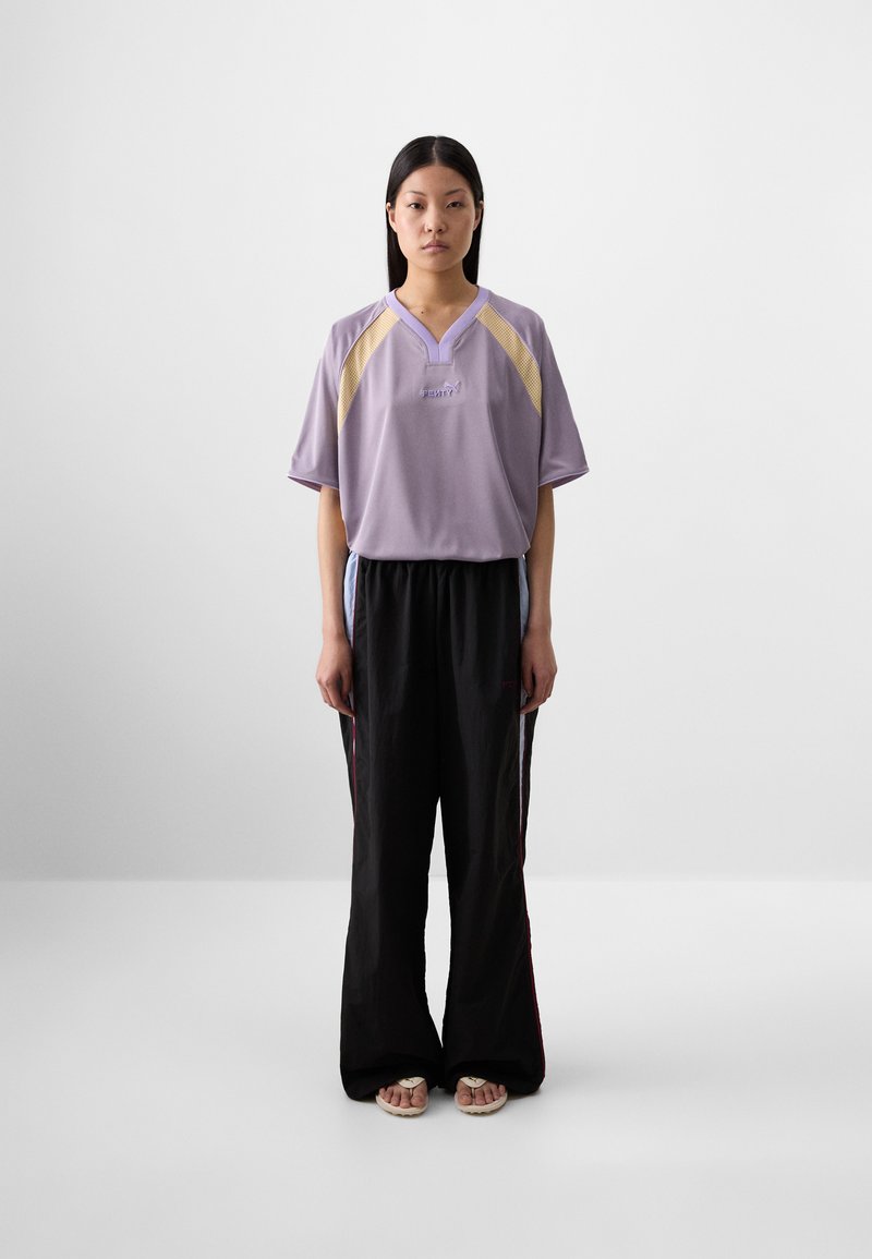 Une jeune femme aux longs cheveux noirs se tient face à l'objectif, portant un t-shirt sportif lavande et un pantalon noir ample, sur un fond uni.