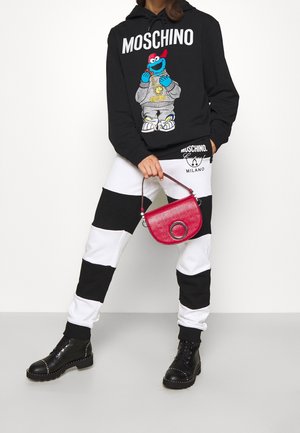 Person, die einen schwarzen Moschino-Hoodie mit Cartoon-Aufdruck und schwarz-weiß gestreifte Hose trägt, eine kleine rote Handtasche hält und schwarze Stiefel trägt.
