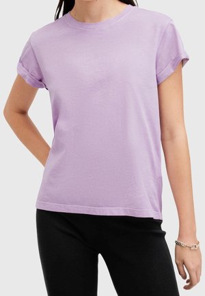 Kobieta ubrana w jasnofioletowy t-shirt z krótkim rękawem i podwiniętymi mankietami oraz czarne spodnie, stoi z jedną ręką na biodrze.