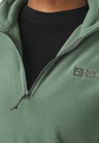 Groene fleece pullover met een half-rits kraag, voorzien van een gestructureerde stof en een klein geborduurd logo op de borst. Zwarte binnenlaag zichtbaar.