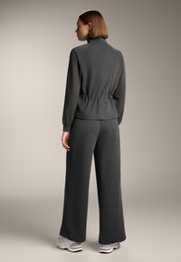 Training set gri cu dungi, având guler înalt, talie elastică și pantaloni evazați. Modelul poartă adidași albi.