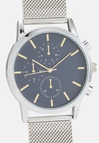 Montre en métal argenté avec un cadran gris foncé, des marqueurs d'heure jaunes et un bracelet en maille. Comprend un sous-cadran et des indicateurs de jour au-dessus du centre.