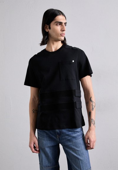 T-shirt noir à manches courtes avec une poche poitrine, arborant trois bandes horizontales noires. Porté avec un short cargo en denim bleu.