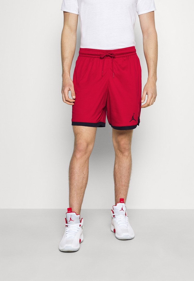 Hombre vistiendo pantalones cortos de baloncesto rojos con ribetes negros y zapatillas altas blancas y rojas, de pie contra un fondo blanco liso.