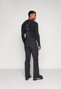 Maglietta nera a maniche lunghe abbinata a pantaloni scuri con bretelle regolabili. L'outfit include scarponi da sci con un marchio laterale.