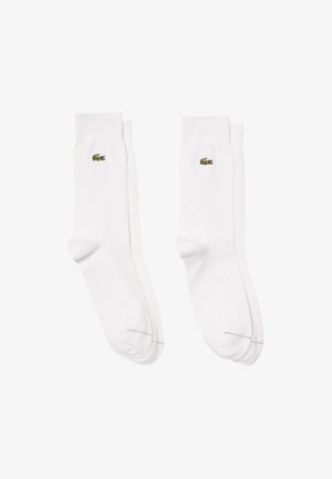 Calcetines de algodón blancos con puños acanalados, que presentan un pequeño logo de cocodrilo verde de Lacoste en el lado exterior. Textura suave y diseño clásico.