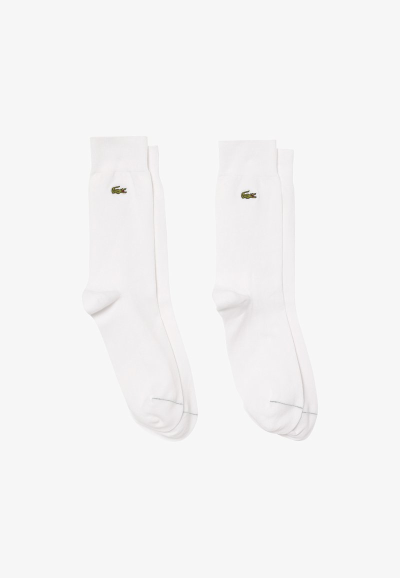 White cotton socks with ribbed cuffs, featuring a small green Lacoste crocodile logo on the outer side. Smooth texture and classic design.