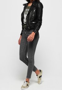 Giacca da motociclista in pelle nera sopra una maglietta con grafiche, abbinata a jeans skinny grigio scuro e sneakers con accenti dorati e strisce nere.