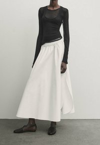Top nero a maniche lunghe con una texture trasparente abbinato a una gonna maxi bianca e fluida con uno spacco laterale, completato da ballerine nere.