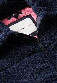 Μπλε σκούρο υφασμάτινο fleece μπουφάν με εν μέρει ανοιχτό φερμουάρ μπροστά που δείχνει ροζ επενδεδυμένη εσωτερική επένδυση και ετικέτα της μάρκας Tommy Hilfiger.
