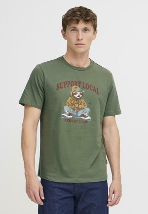 T-shirt en coton vert olive avec un paresseux imprimé portant un sweat à capuche et le texte "SOUTENEZ LES CHATS DE RUES LOCAUX." Manches courtes, design à col rond.