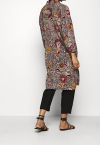 Kobieta nosząca płaszcz w kwiatowy wzór do kolan z podwiniętymi rękawami, czarne spodenki 3/4 oraz czarne płaskie sandały na paski.