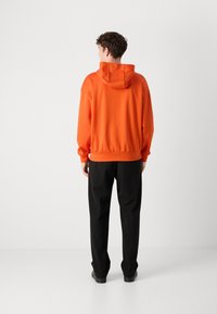 Felpa arancione con cappuccio regolabile, polsini e orlo a costine, abbinata a pantaloni neri. Tessuto morbido, vestibilità comoda, design senza cuciture.