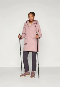 Jachetă waterproof de culoare roz deschis, cu glugă și buzunare frontale, purtată peste pantaloni negri și pantofi sport, ținând bețe de schi într-un cadru neutru.