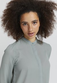 Blusa verde chiaro, con bottoni, colletto alto, maniche lunghe e una texture liscia. Caratterizzata da un design minimalista e una morbida caduta.
