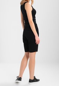 Haut sans manches noir assorti à un short noir. Modèle portant des baskets noires avec des accents cloutés. Fond neutre.