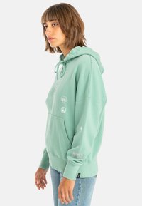 Mint green hooded sweatshirt with a kangaroo pocket, featuring various small graphics and text on sleeves. Soft cotton blend fabric.
