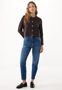 Hnědá zkrácená bunda s límcem, zapínáním na knoflíky a dvěma kapsami, kombinovaná s modrými zúženými džíny a černými mokasíny.