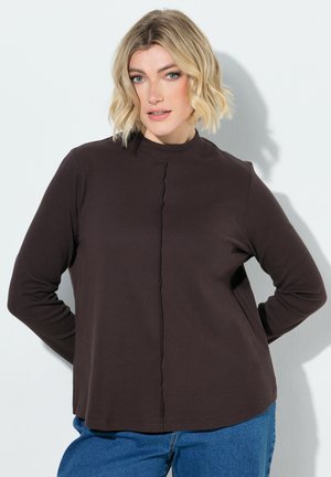 Blonde vrouw met golvend haar, dragend een donkerbruin topje met lange mouwen en een blauwe spijkerbroek, staand met de handen achter haar rug.