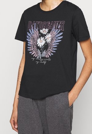 Personne portant un t-shirt noir avec le texte "DAYDREAMER", des fleurs blanches, un graphique d'ailes violettes et un texte en cursive "my energy creates my reality."