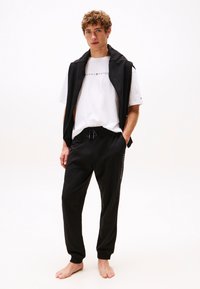 Fekete melegítőnadrág elasztikus mandzsettákkal, fehér logós pólóval párosítva. A modell fekete ujjatlan dzsekit visel.