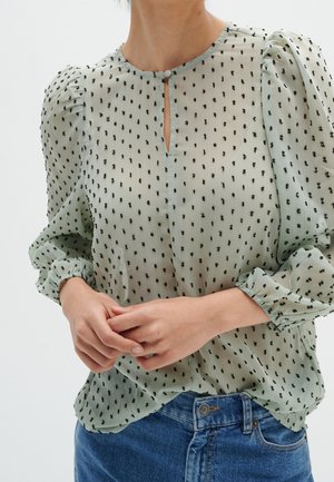 Mujer con blusa transparente de color verde claro con lunares negros, mangas abullonadas y escote en forma de ojo de cerradura, combinada con vaqueros azules.
