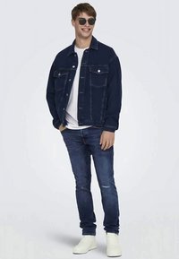 Chaqueta de mezclilla azul oscura con dos bolsillos en el pecho, camiseta blanca, jeans azul oscuro y zapatillas blancas; textura suave, corte informal.