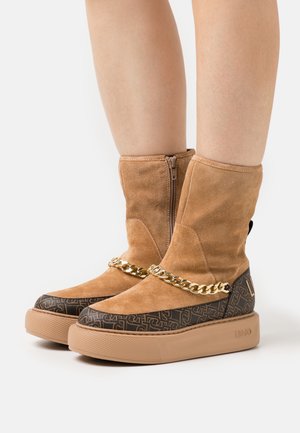 Beige mockaboots till ankeln med guldkedjeaccent, med ett mönstrat svart nedre parti, låg gummisula och dragkedja baktill.