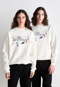 Sweat-shirts crème avec un imprimé graphique comportant des éclaboussures de couleur et du texte, portés avec un pantalon noir ; coupe décontractée, poignets côtelés.